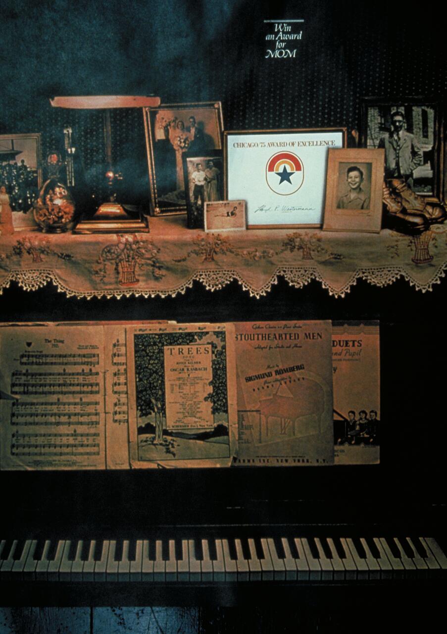 A vintage piano with sheet music on the stand, framed family photos, and an award above the keys. A decorative lace cloth holds additional memorabilia, adding a nostalgic atmosphere.