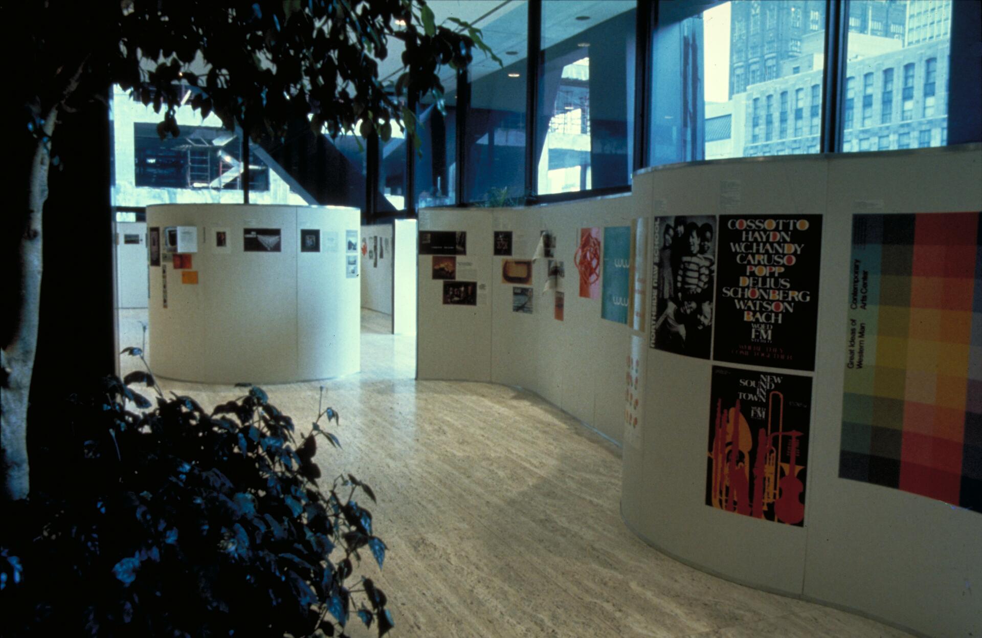 Curved indoor gallery space with posters displayed on white walls. Large windows in the background show a view of buildings outside. A leafy plant is visible in the foreground.