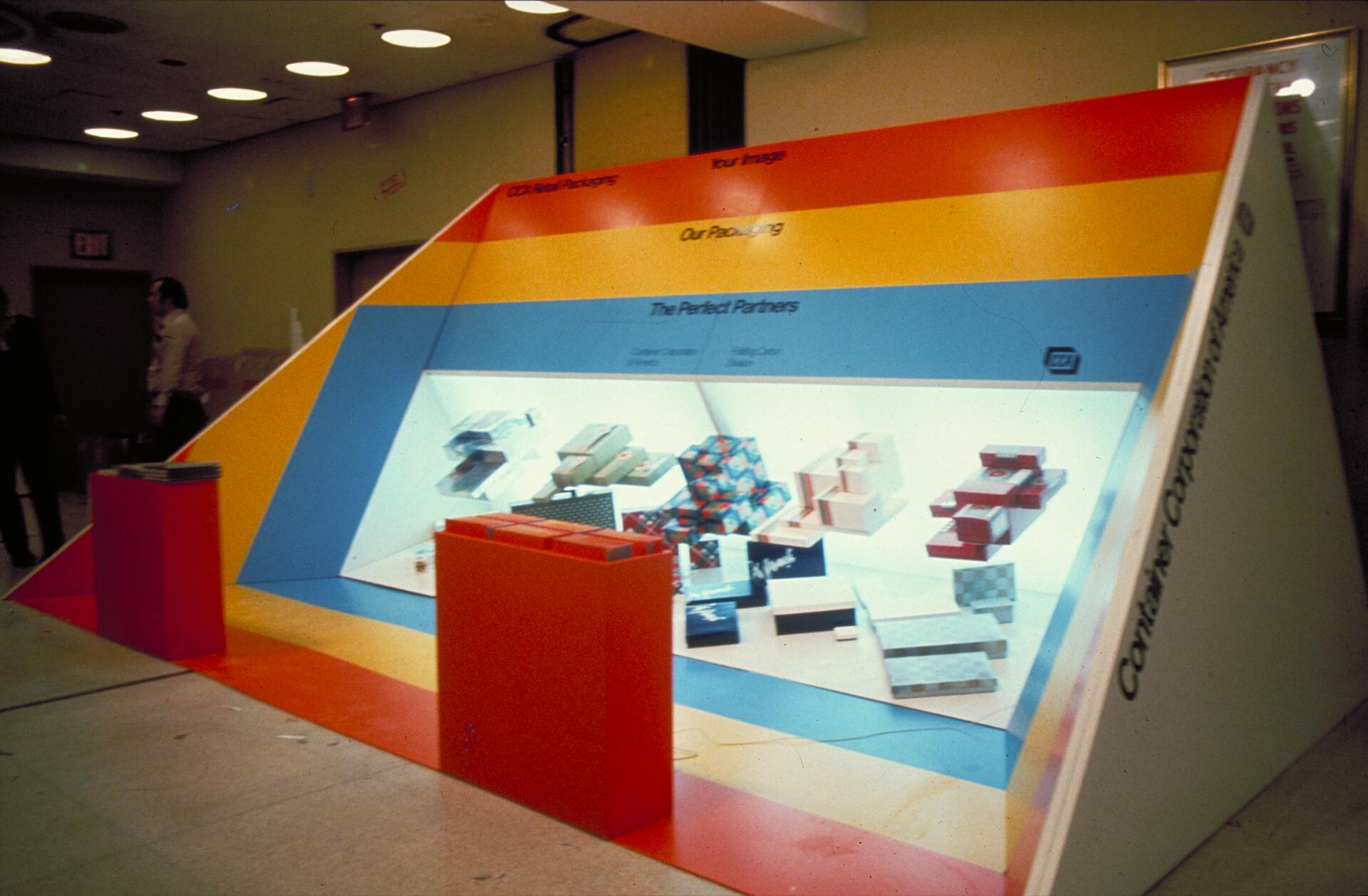 Colorful exhibition booth displaying various packaging boxes, with gradients of red, orange, yellow, and blue, and an illuminated showcase. Two people stand nearby in a dimly lit room.
