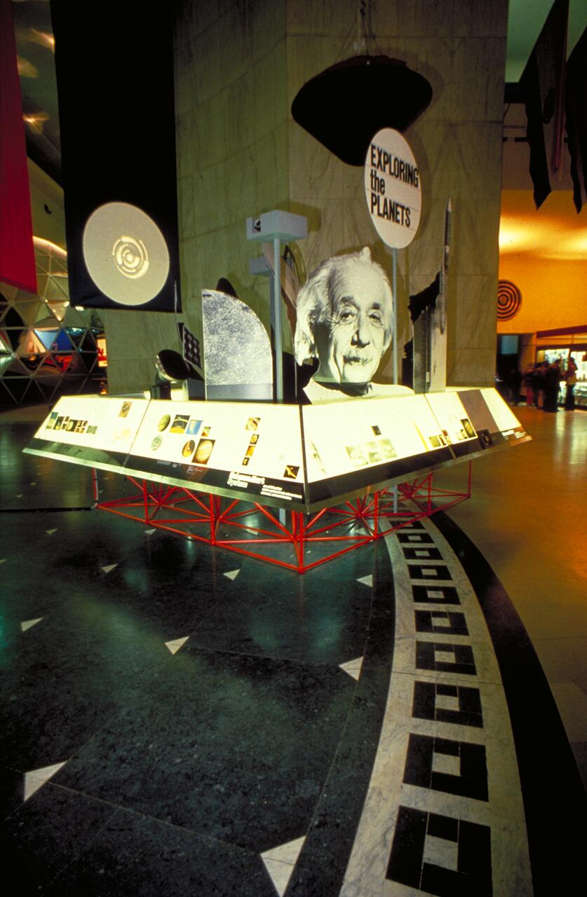 Science exhibit featuring a large portrait, informational panels, and display stands under dim lighting, with a sign reading "Exploring the Planets."