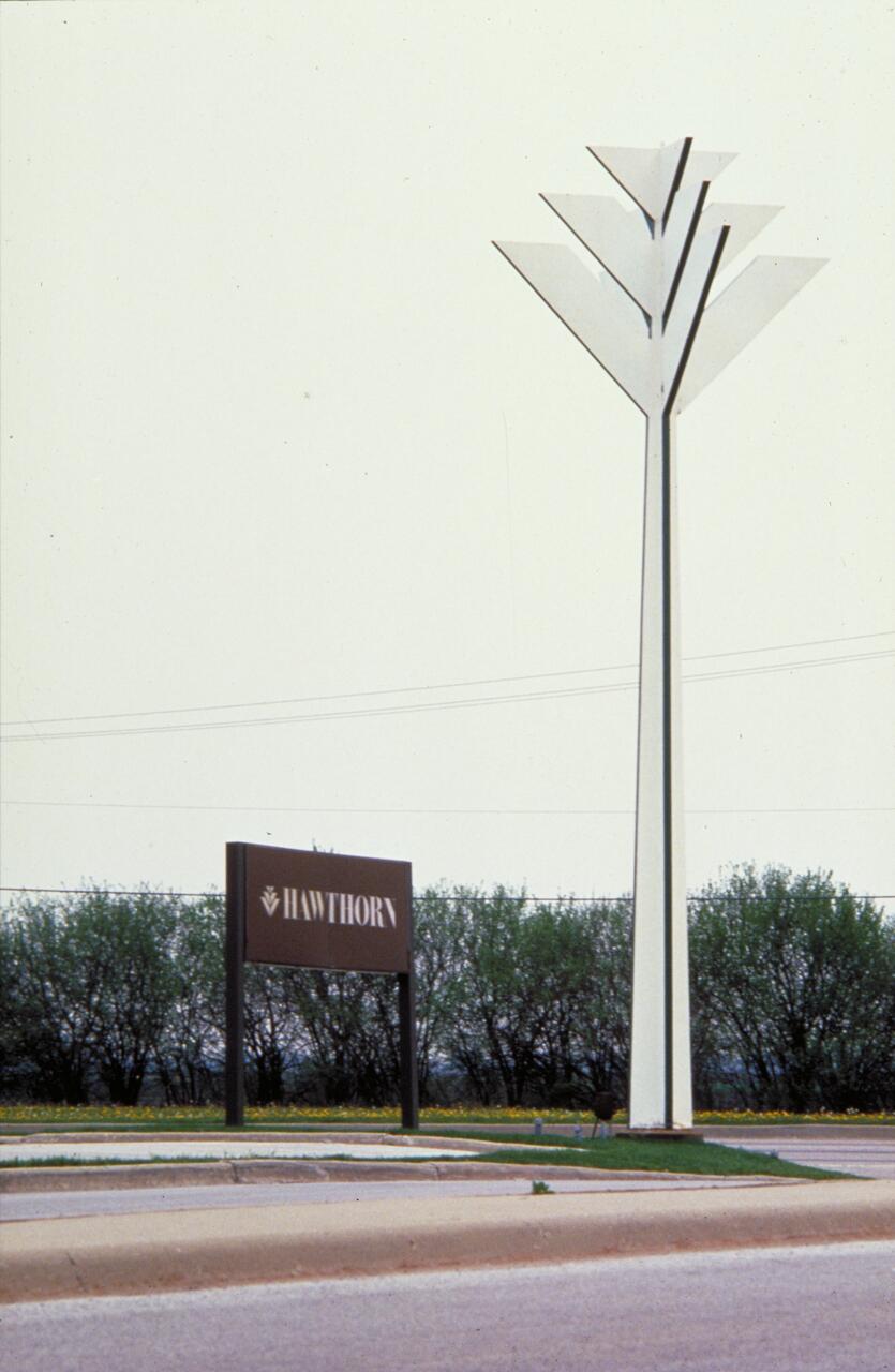 Tall metal sculpture resembling a tree beside a "Hawthorn" sign, set against a backdrop of bushes and a clear sky.