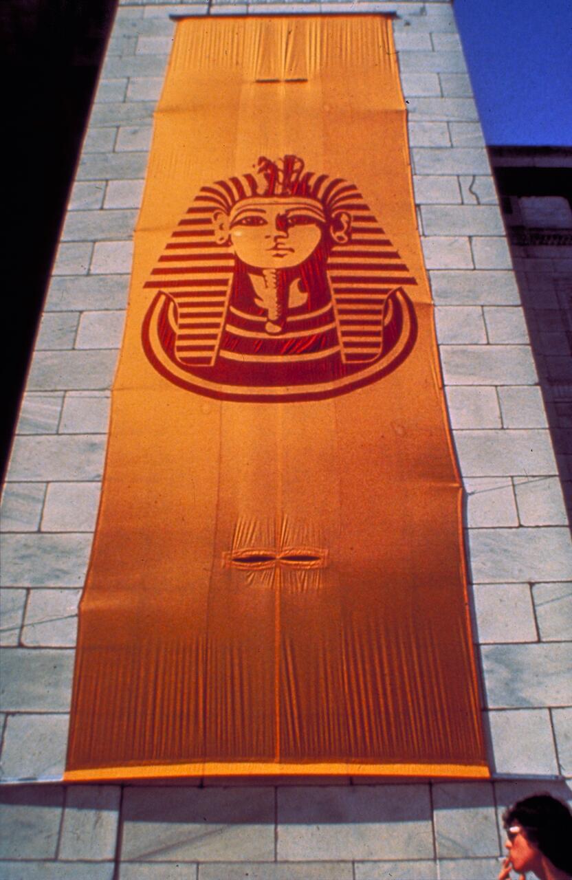 Large orange banner on a stone wall featuring an Egyptian pharaoh's head in brown. A person with dark hair stands at the bottom right, looking up. The sky is clear blue.