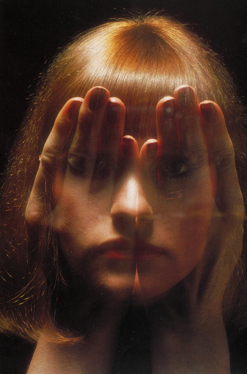 A woman with reddish hair appears with her face partly distorted, hands covering her eyes, creating a mirrored, surreal effect.
