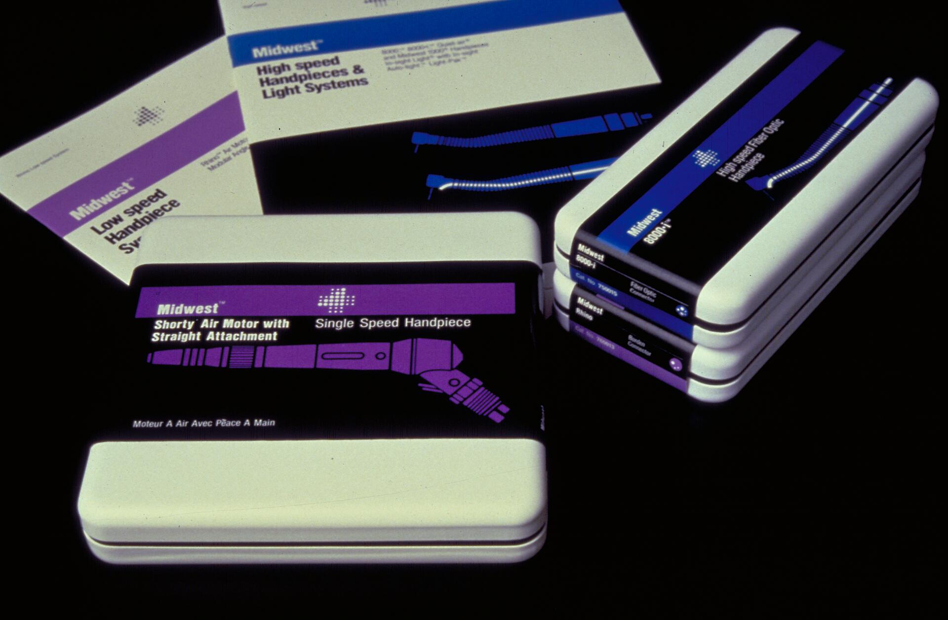 Stacked boxes of dental handpieces featuring black, blue, and purple labels with text and images of dental tools. White sheets with matching labels lie on top. Black background.