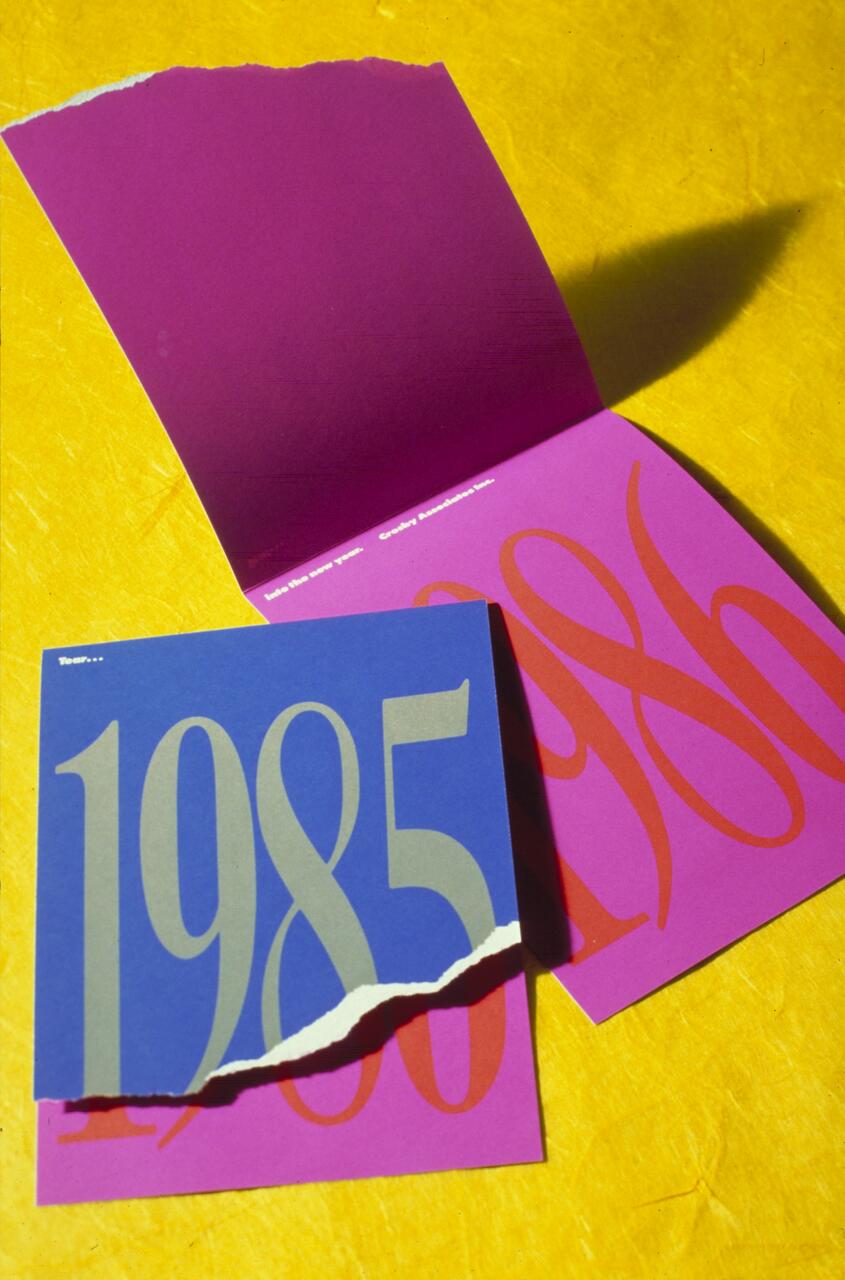 Folded purple and blue cards on a yellow background, displaying large numbers "1985" in gray on blue and "1986" in orange on purple. A torn edge divides the years.