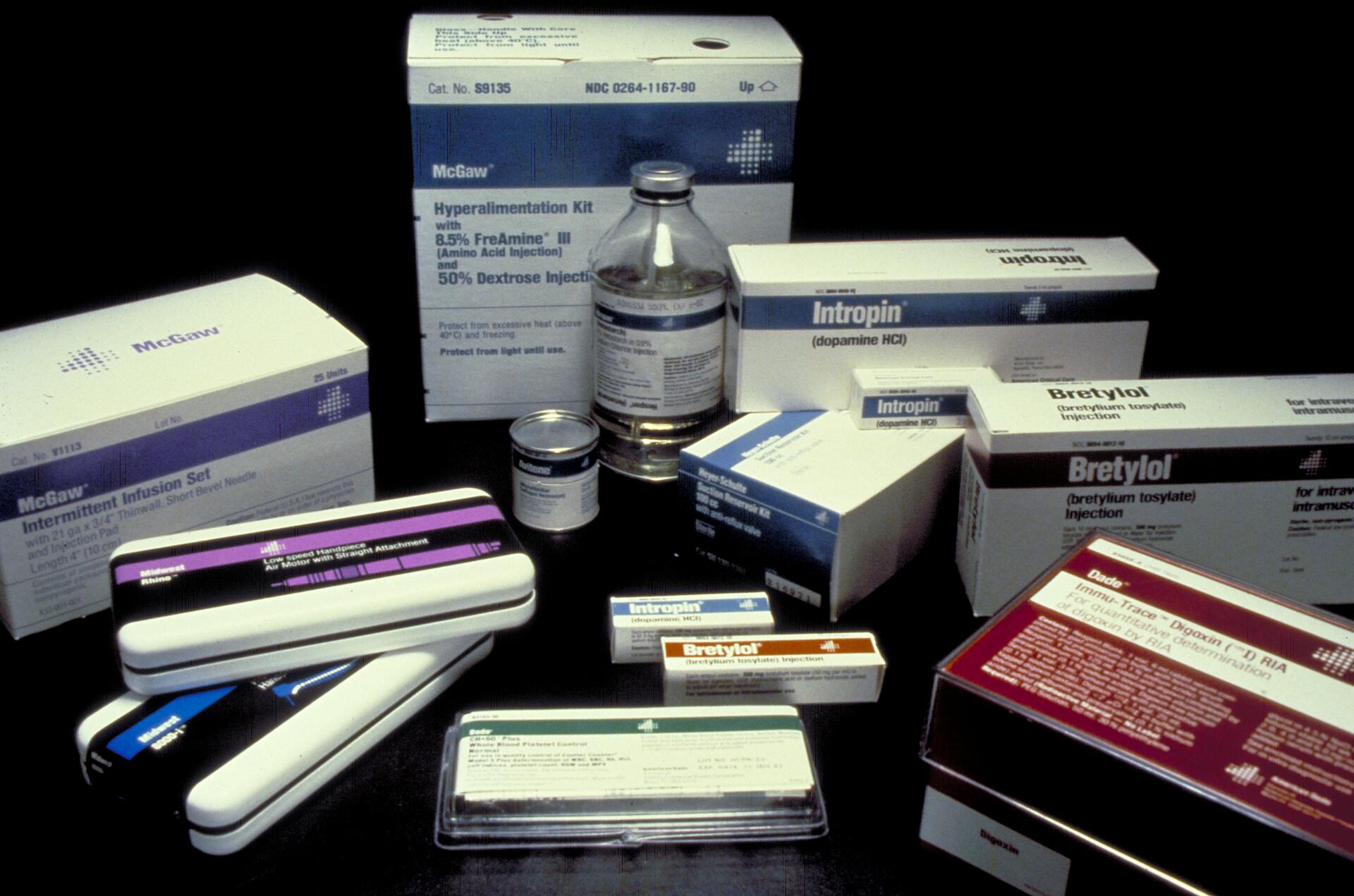Various medical supply boxes and vials arranged on a dark surface, including packages labeled Bretylol, Intropin, and a hyperalimentation kit.
