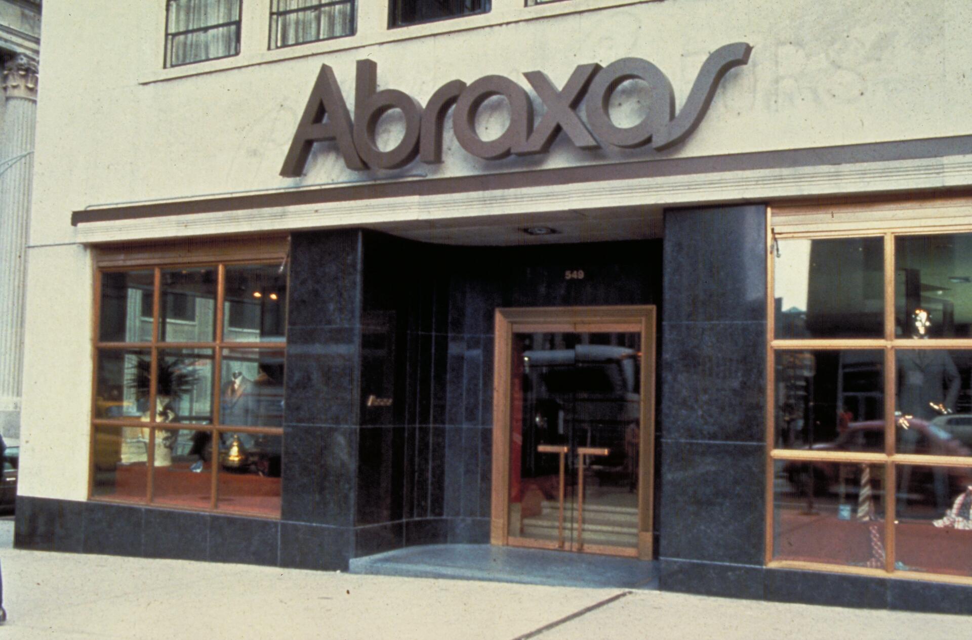 Storefront with "Abraxas" sign in large letters above the entrance. The facade features dark panels and large glass windows displaying mannequins. Double glass doors with wooden frames lead inside.