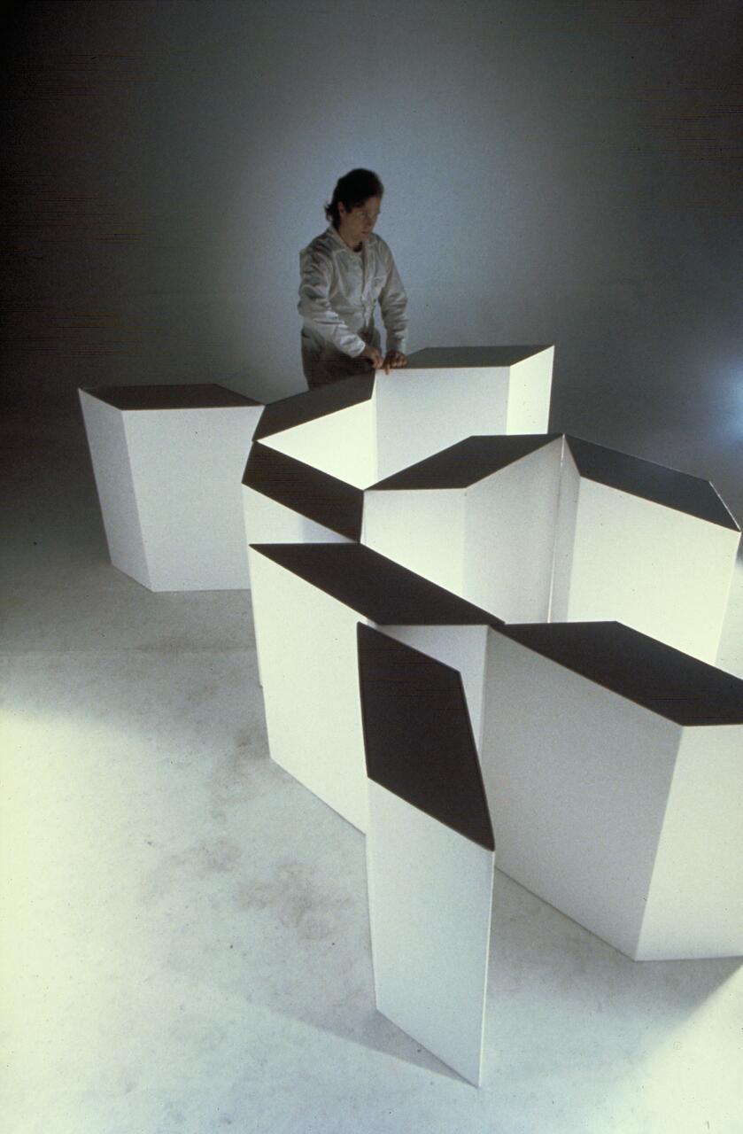 A person in a light-colored outfit interacting with a geometric, abstract white sculpture composed of interlocking, angular shapes on a neutral background.