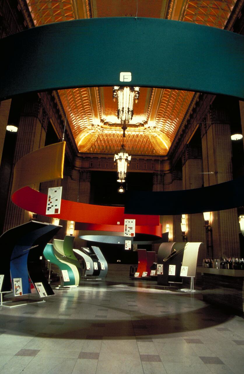 Exhibition hall with colorful, curved panels suspended from the ceiling. The ornate ceiling features geometric patterns, illuminated by chandeliers. Information boards are displayed on some panels.