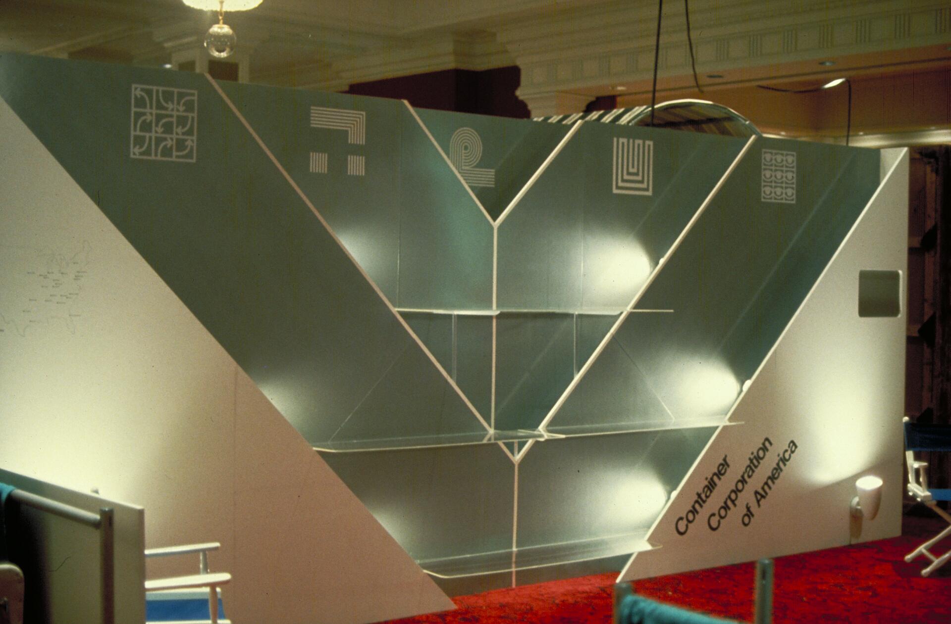 Trade show booth for Container Corporation of America with a geometric design. The wall features various abstract symbols and has illuminated shelving. The setting includes red carpet flooring and a chandelier above.