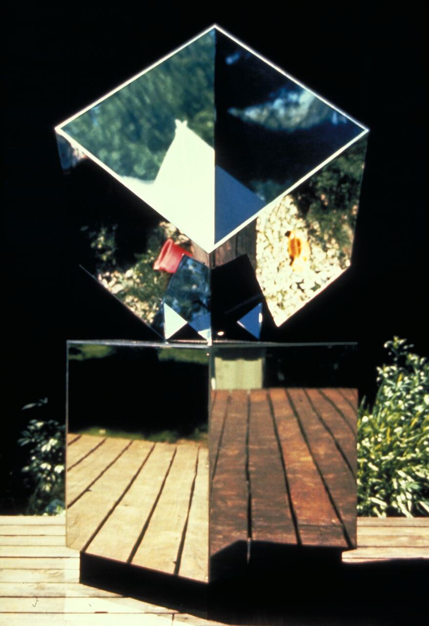 Reflective sculpture of two stacked mirrored cubes on a wooden deck, displaying varied reflections of the surrounding greenery and forest, with a high contrast between the bright reflections and the dark background.