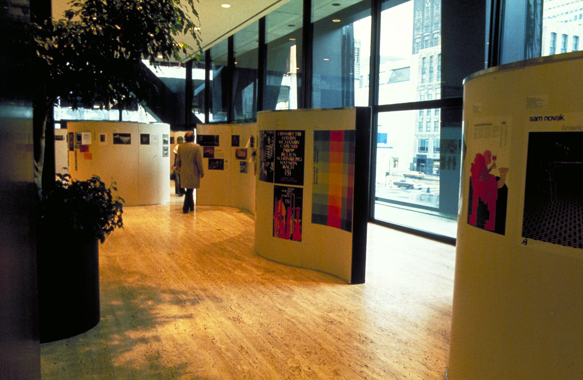 Art exhibition in a bright, modern gallery with tall windows. A person walks through curved displays featuring colorful graphics and posters. Indoor plants and cityscape visible through the windows.