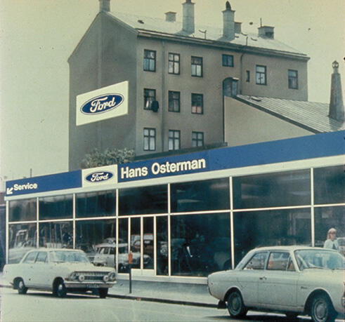 Ford dealership in an urban setting with vintage cars parked in front. The building has a blue sign with "Hans Osterman" on it, and a Ford logo is visible on the wall. A multi-story building is in the background.