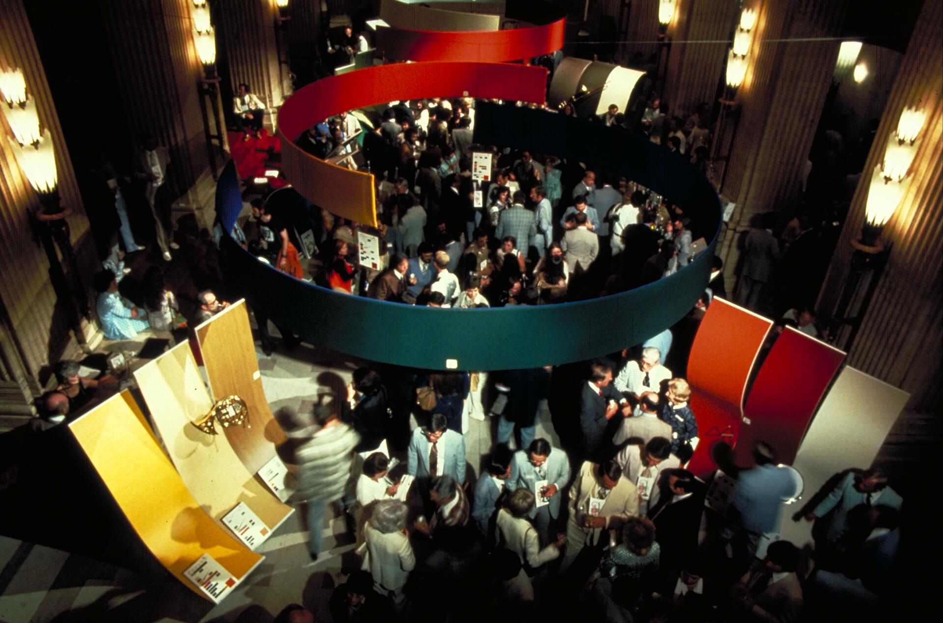 Crowded exhibition hall with people mingling around colorful, curved panels suspended from the ceiling. Warm lighting creates a lively atmosphere.