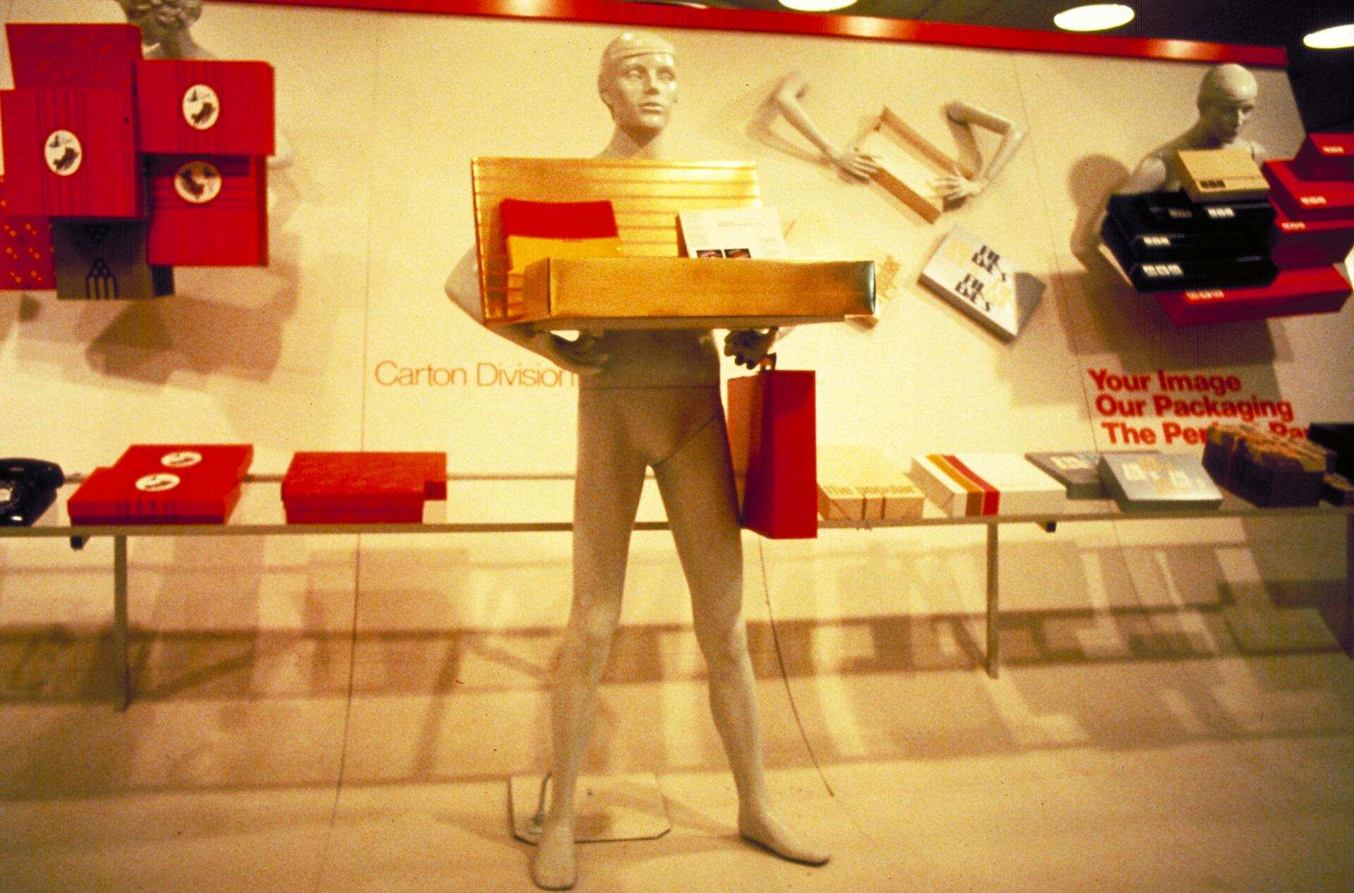 Mannequin holding packaging samples, surrounded by red and black boxes on display shelves.