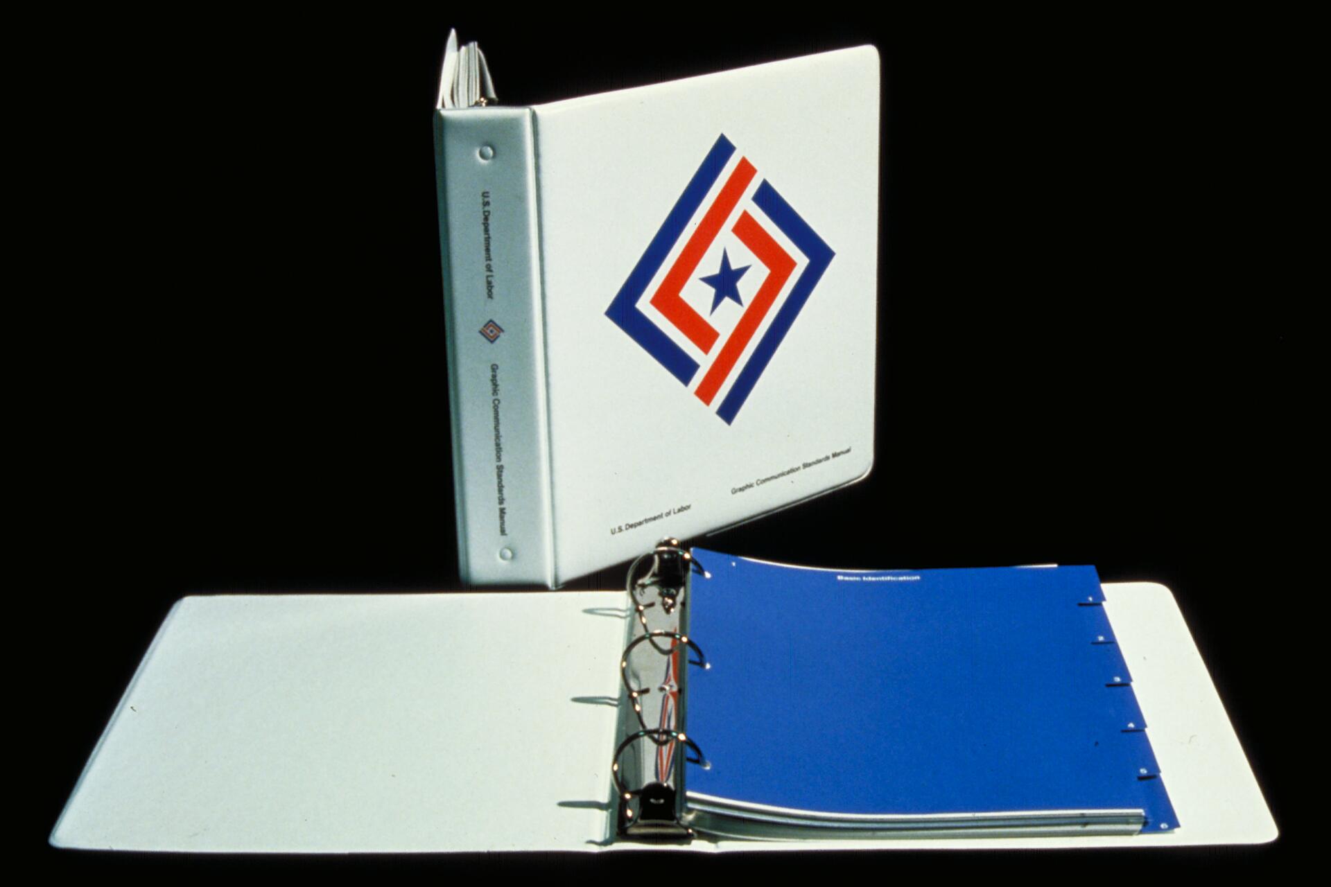 Two white binders with colorful graphic design on the front, one standing open with blue dividers visible, on a black background. The text on the binder references the U.S. Department of Labor.