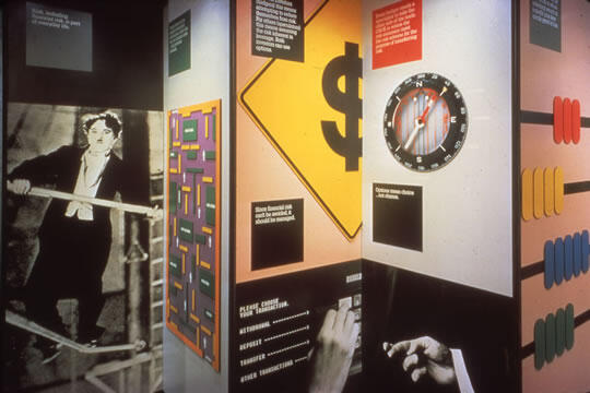 Exhibit display with a black-and-white photo of a person balancing, a dollar sign on a yellow background, a circular clock, and colorful abacus beads.