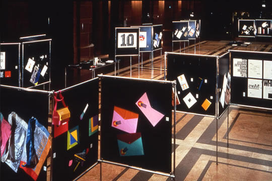 Art exhibition with various black panels displaying colorful abstract graphics and designs, set in a dimly lit gallery space. The floor is patterned with light and dark tiles.