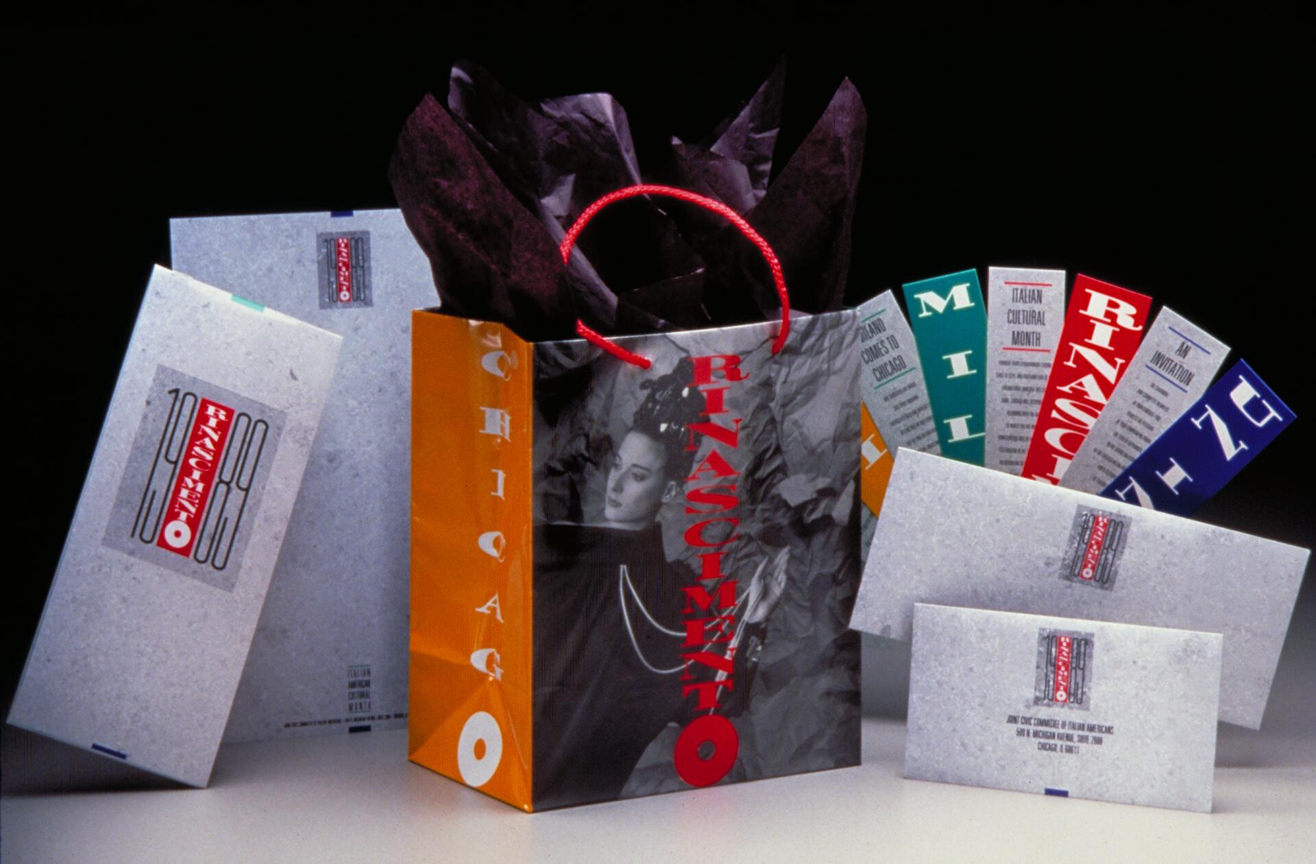 Shopping bag and envelopes with modern graphics, including a black-and-white portrait and the word "Rinascimento" in bold red. A red handle and dark tissue paper emerge from the bag, surrounded by colorful brochures.