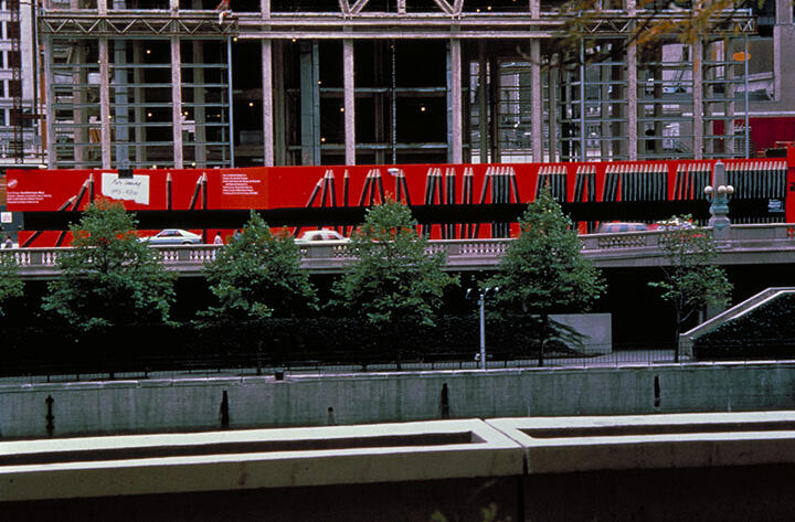 Construction site with steel girders and a bright red barrier featuring geometric designs. Cars are visible behind a row of trees, and a partially built structure is in the background.