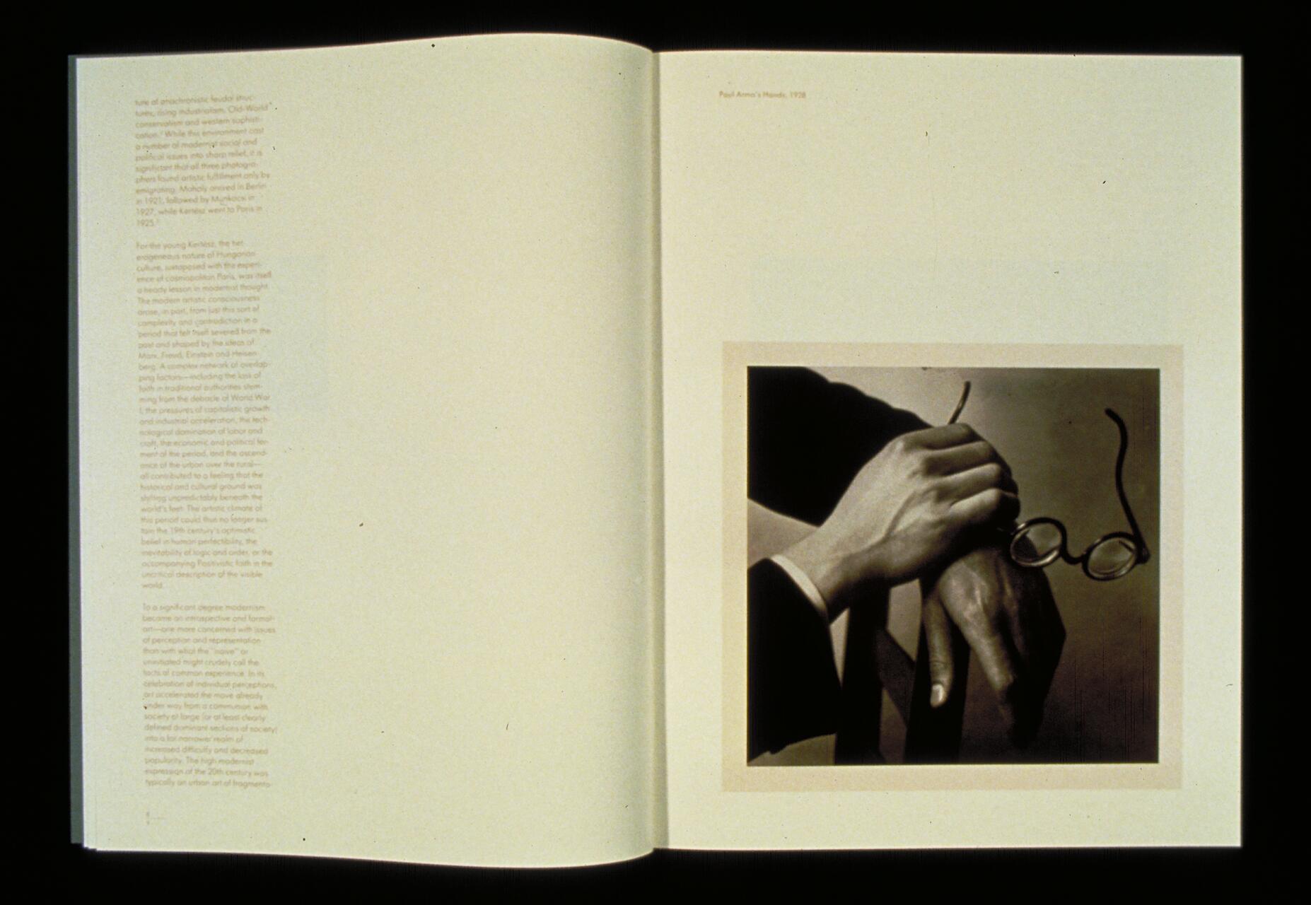 Open book displaying a sepia photograph of two hands. One hand holds a pair of glasses, while the other rests on a surface. Text is visible on the left page.