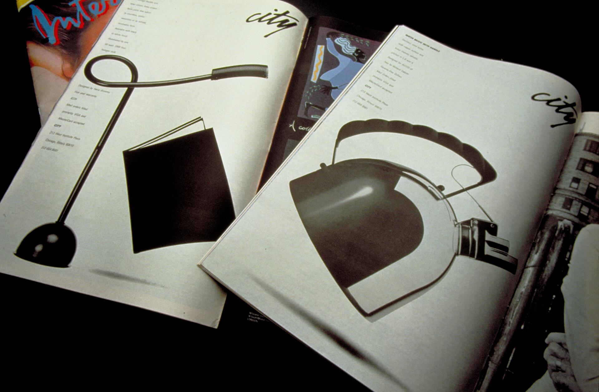 Open magazine showing pages with minimalist product designs; one page features a sleek lamp with a looped neck, the other a modern kettle with a curved handle. Both pages have the word "city" in stylized text.