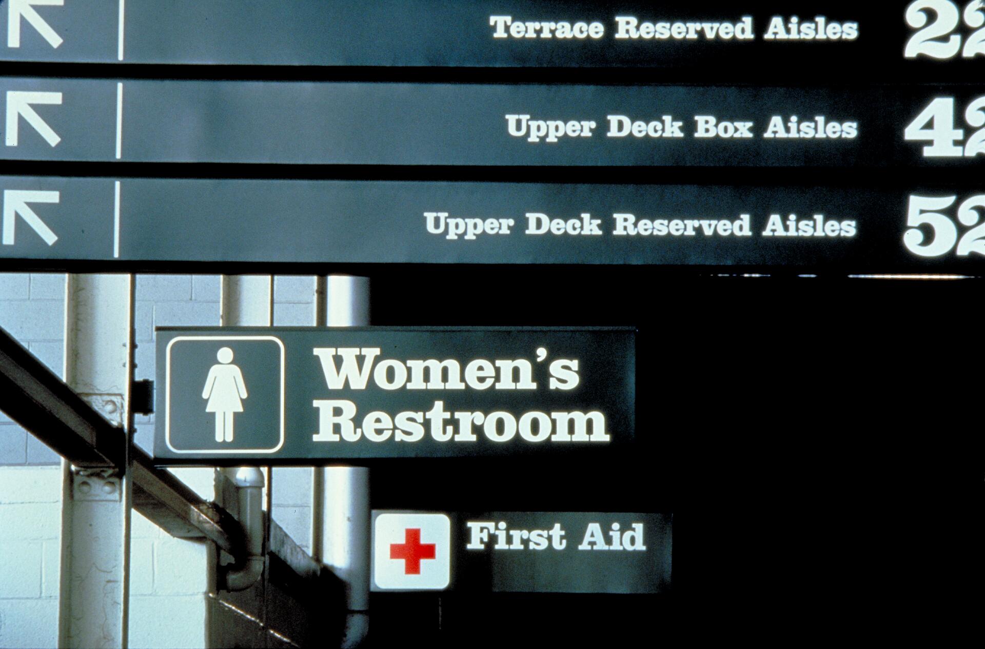 Directional signs in an indoor venue. Arrows point left to various aisle sections. Prominent signs for Women's Restroom and First Aid.