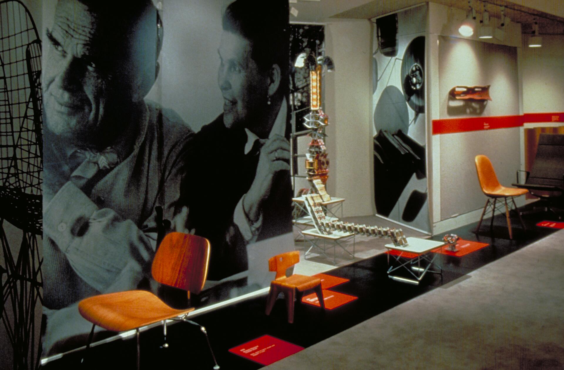 Exhibition space featuring modern chairs and architectural models on display, with two large black-and-white portraits in the background.