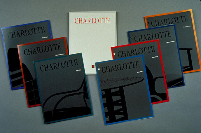 Eight brochures titled "Charlotte" arranged in a semicircle on a gray surface. Each cover features black silhouettes of furniture, framed in different colors: blue, red, orange, and green. The central brochure is white with red text.