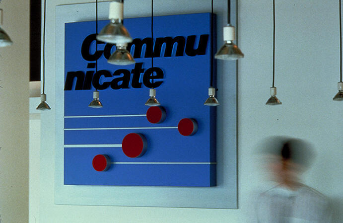Blue wall art with the word "Communicate" and red dots on horizontal lines, surrounded by hanging lights. A blurred figure walks by.