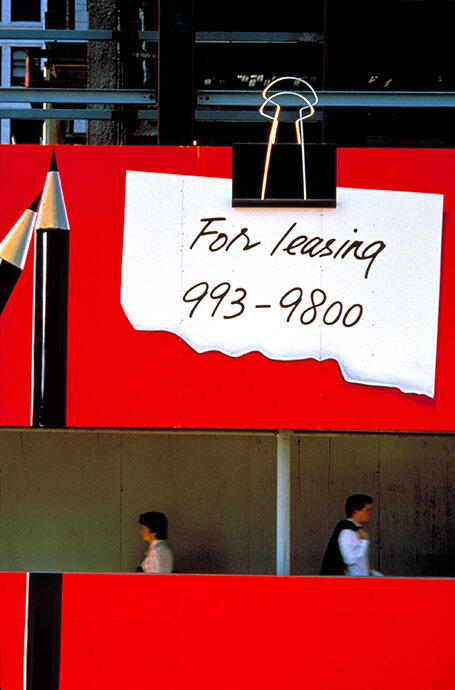 Red wall with a large note clipped, featuring bold text. A black and silver pencil is on the left, while an empty walkway is below, with two blurred pedestrians passing by.