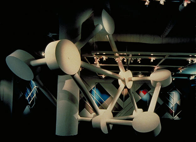 Sculptural installation with large white geometric shapes connected by rods, illuminated by overhead lights in a gallery setting, with abstract art on the walls in the background.