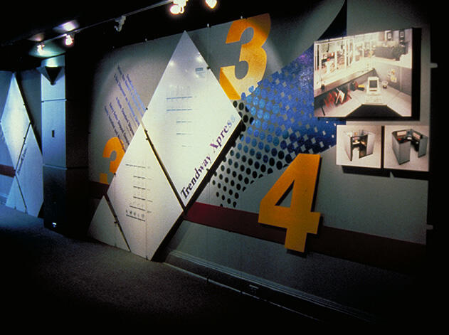 Exhibition wall with large yellow numbers 3 and 4, geometric panels, and small framed photographs. Dim lighting with focused spotlights.