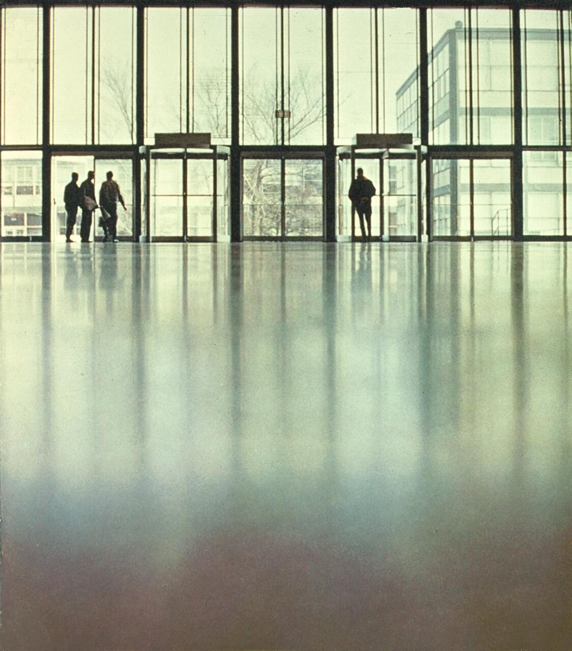 Spacious, empty hall with a glossy floor, large glass windows, and a few silhouetted figures near the entrance.