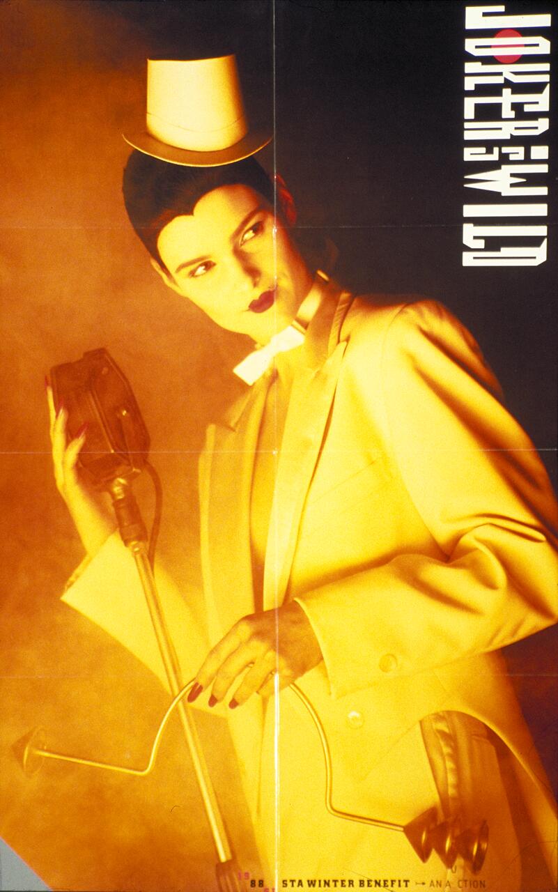 Person in a yellow blazer and white top hat holds a vintage microphone. Their hair is styled neatly, and they have dark lipstick. The background is a warm tone with abstract text on the side.