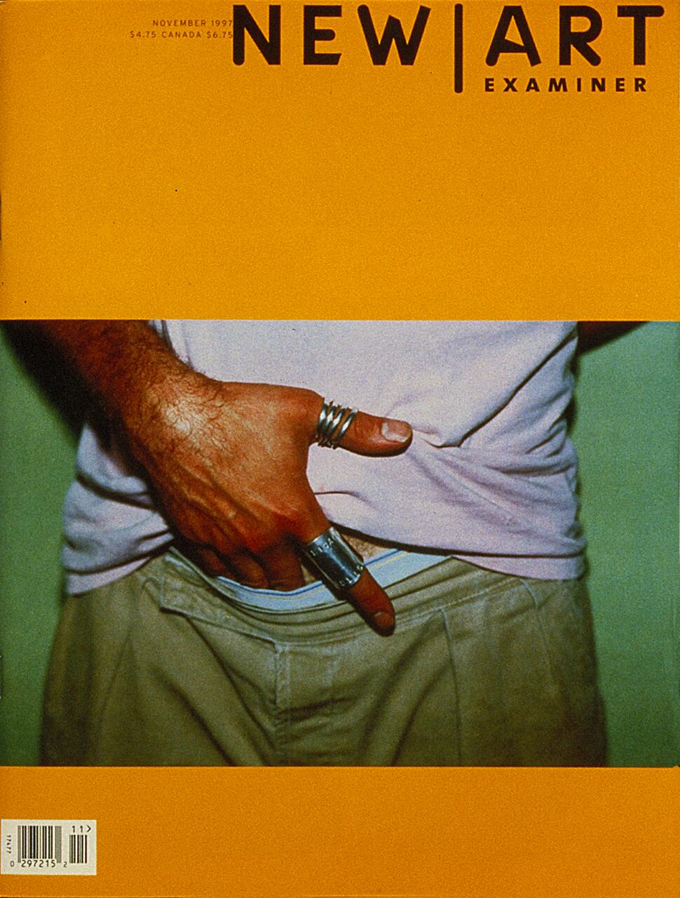 Magazine cover with a close-up photo of a man's hand wearing silver rings, tucking a thumb into khaki pants. The top of the cover is bright orange with the title "NEW ART EXAMINER."